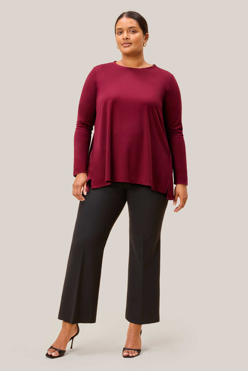 Model wearing plus size burgundy jersey top with dipped hem and side slits, styled with tailored trousers. THE HOUR