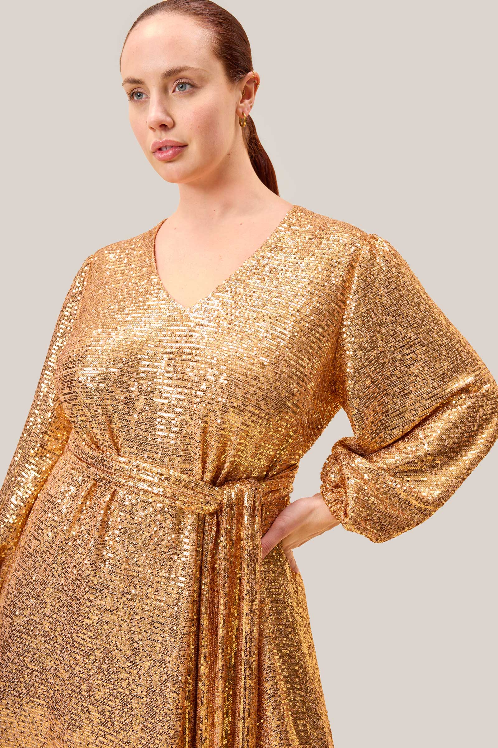 Close-up of a plus-size model in a gold sequin gown showing the self-tie waist belt and blouson sleeves for an elegant silhouette. THE HOUR