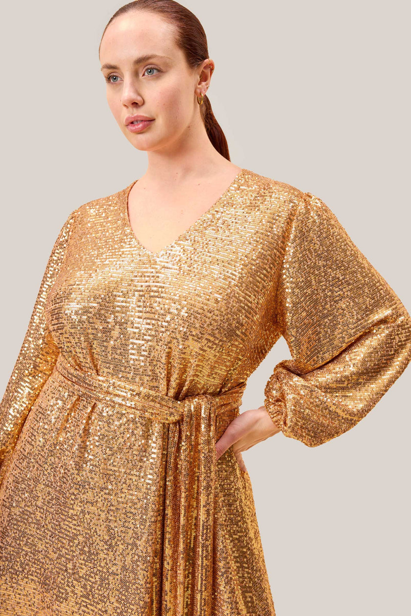 Close-up of a plus-size model in a gold sequin gown showing the self-tie waist belt and blouson sleeves for an elegant silhouette. THE HOUR