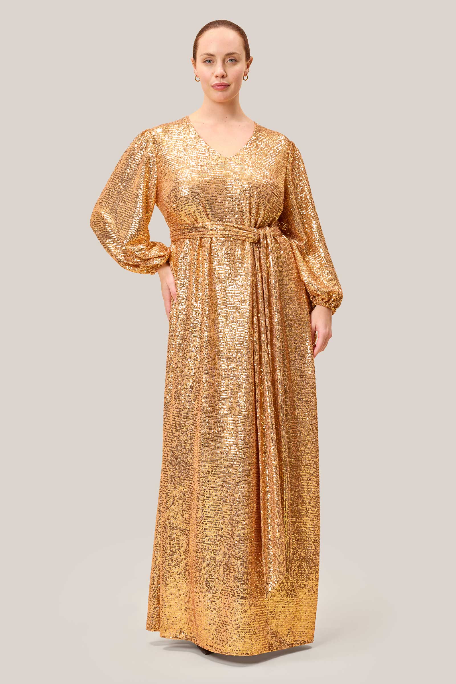 Plus-size model wearing a floor-length gold sequin gown with long sleeves and a V-neckline, designed by The Hour