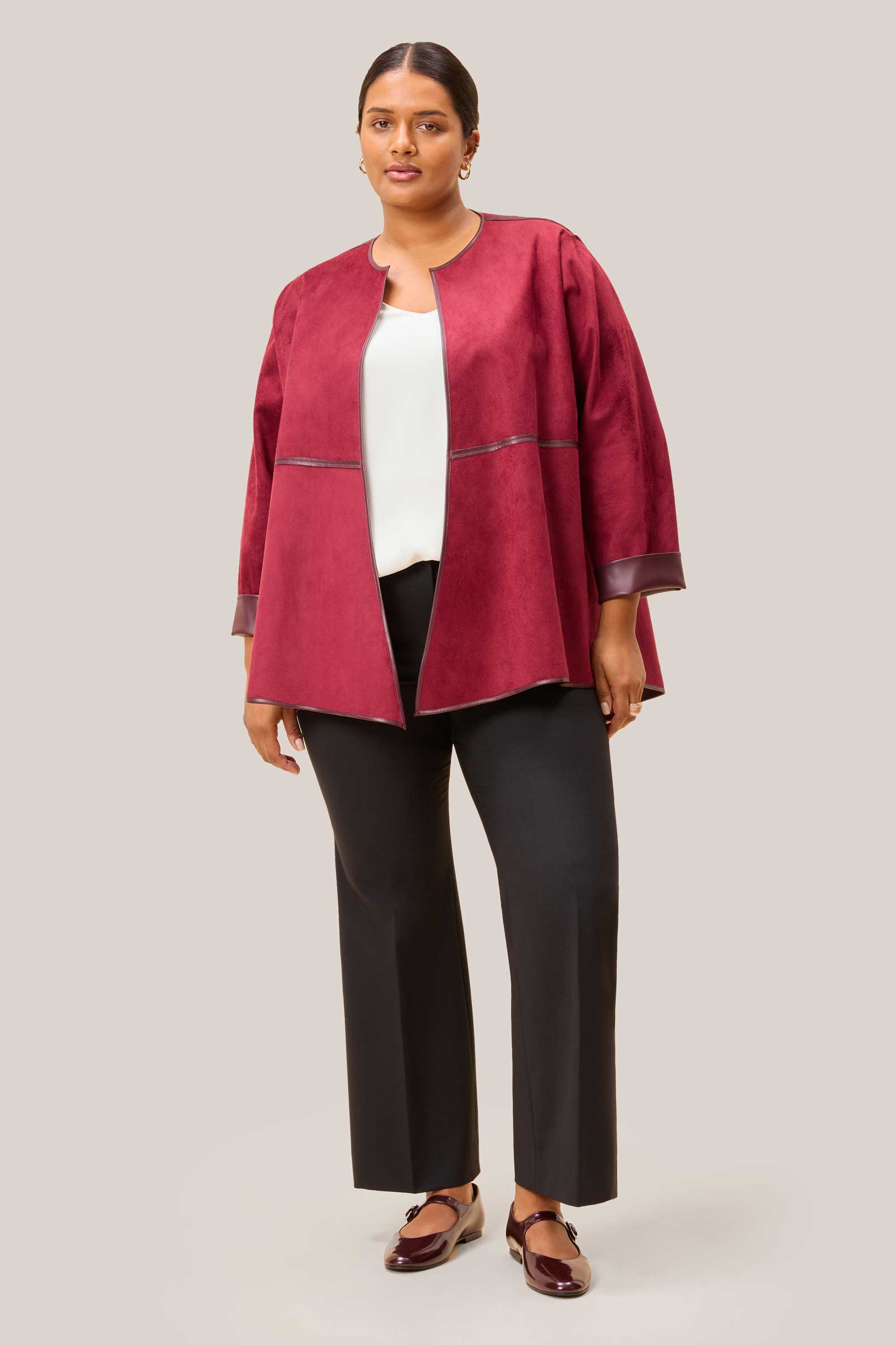 Plus-size woman wearing a burgundy suede jacket over a white top and black pants on a light grey background