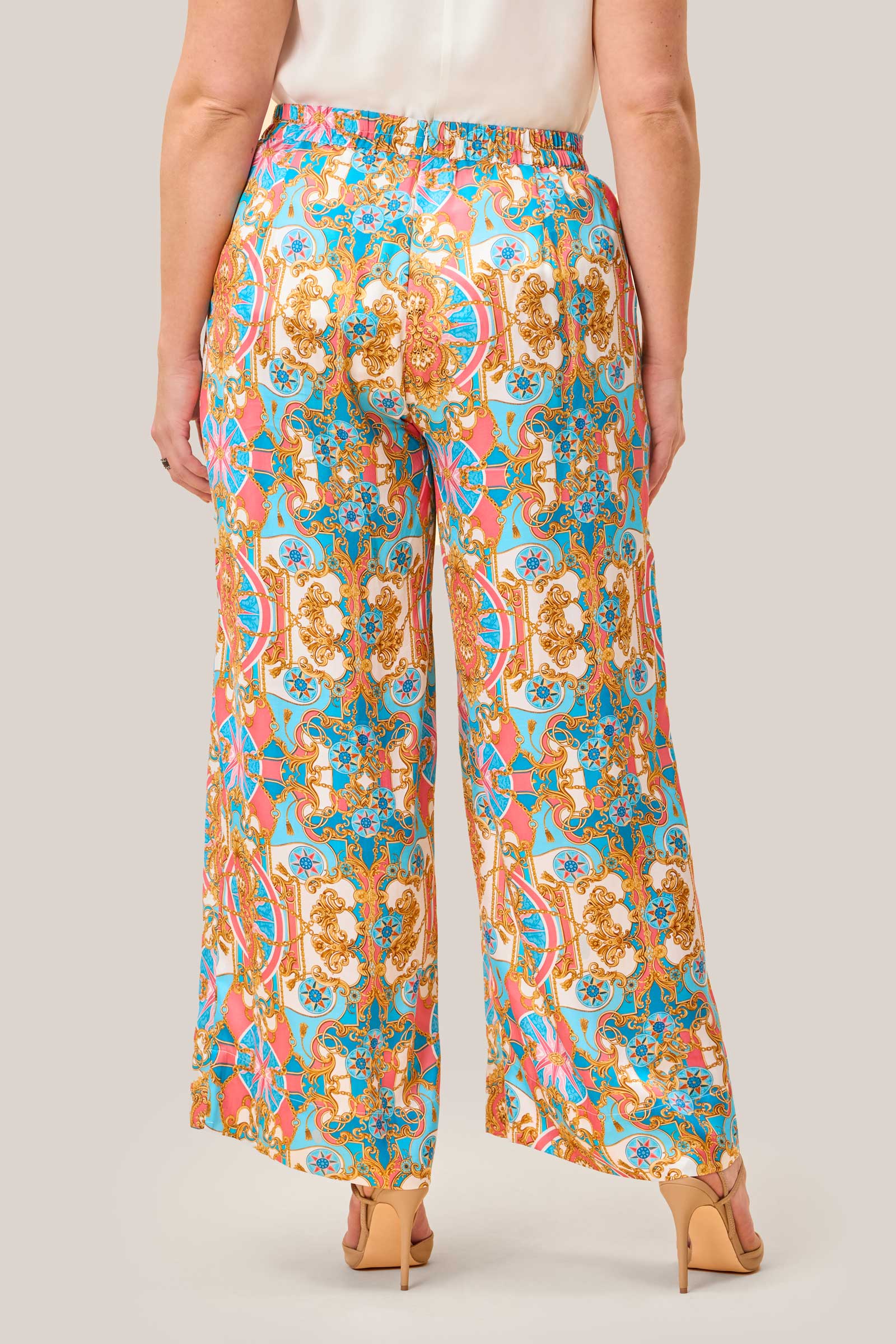 Back view of wide leg satin palazzo trousers in baroque print with gold, coral pink, and aqua blue design, shown on plus size model wearing nude heels.