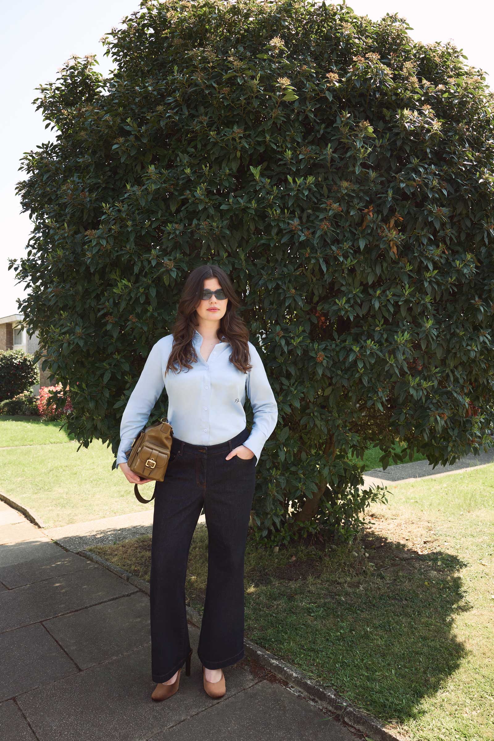 Plus size model wearing dark indigo high waisted wide leg jeans and light blue shirt from THE HOUR, styled with heels and a tan Chloe camera bag, standing outdoors in street style look.