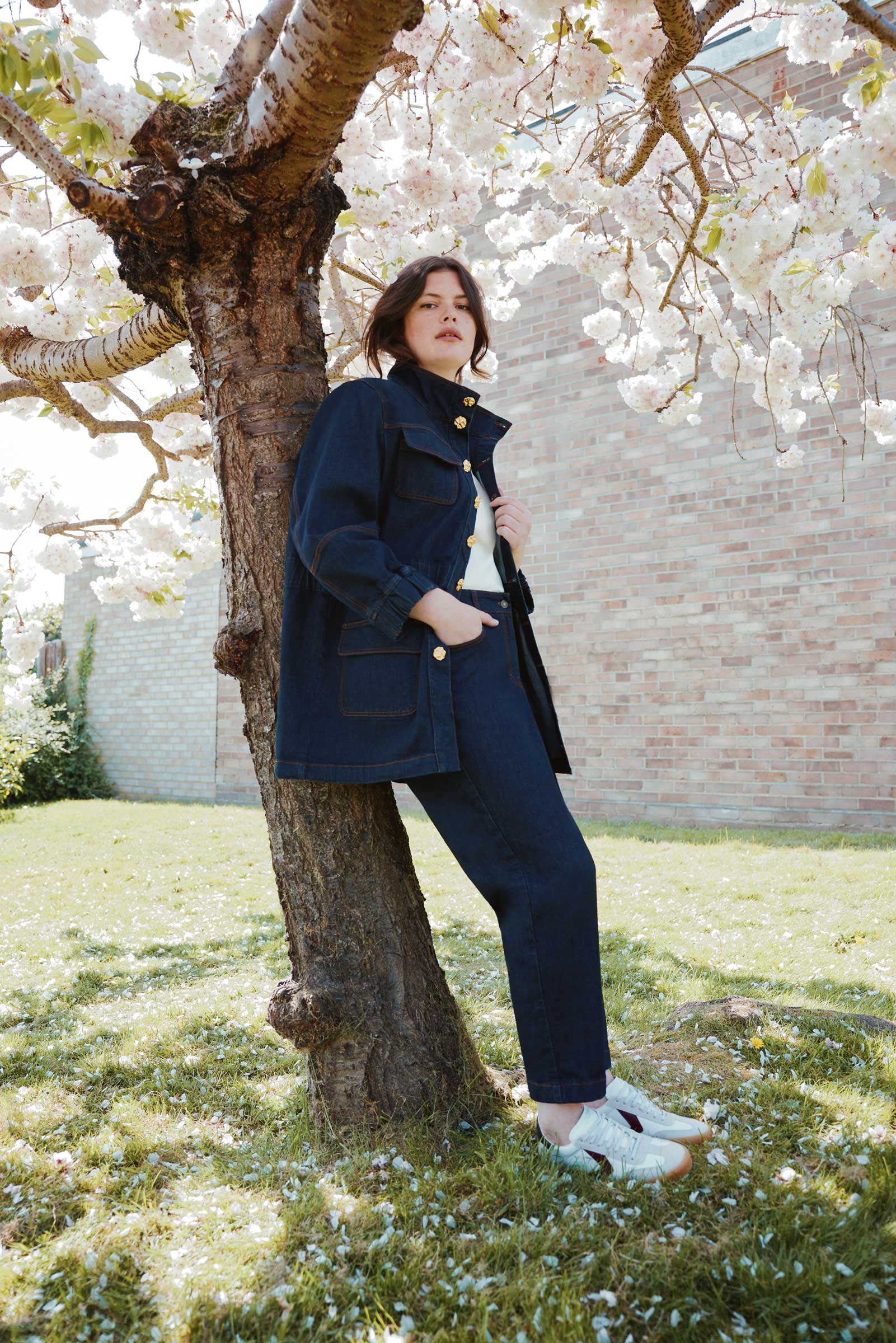 Plus size model wearing a dark indigo denim jacket and tapered jeans from The Hour, leaning against a tree in spring blossom setting.
