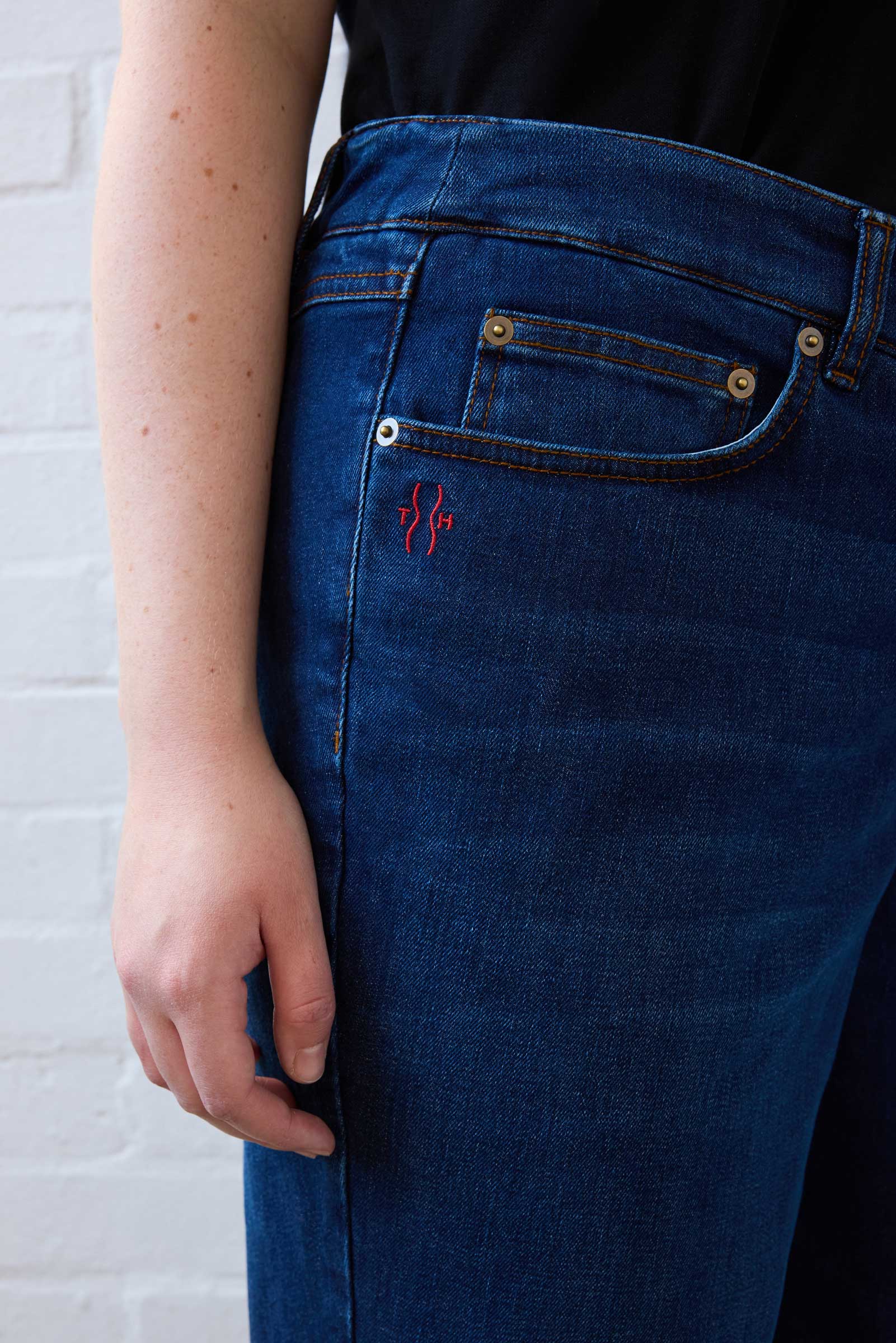 Detail of red TH logo embroidery above front pocket on mid-blue jeans