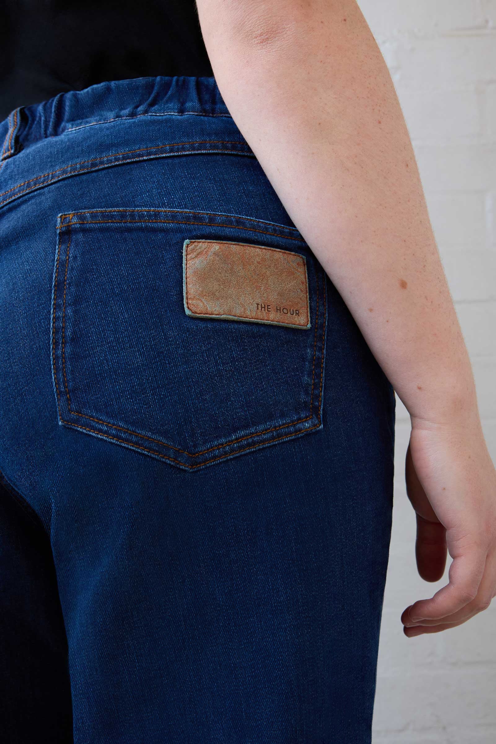 Close-up of branded leather patch on back pocket of mid-blue tapered jeans. THE HOUR