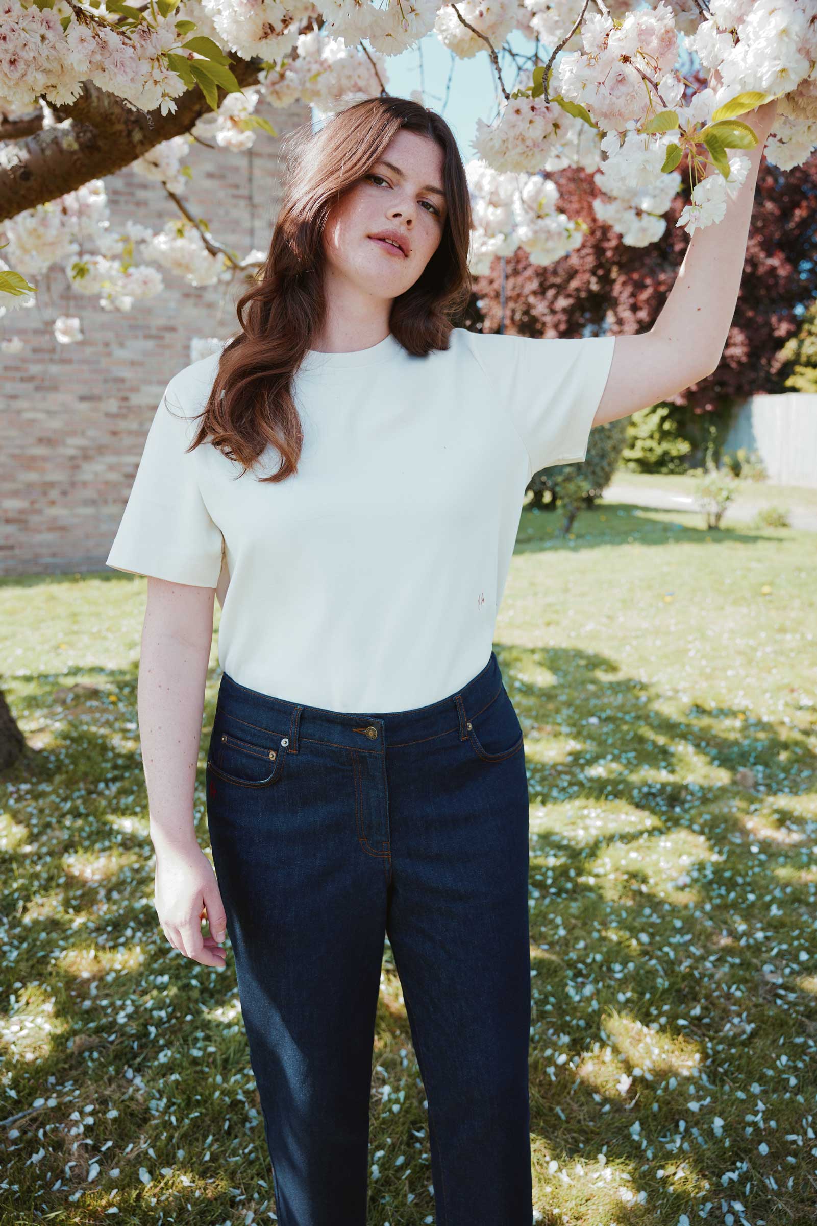 Plus size model wearing cream organic cotton T-shirt and dark denim jeans, standing under blossoming tree by The Hour