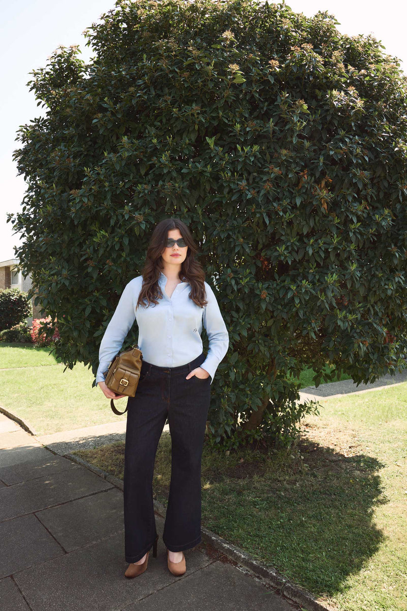 Plus size model wearing dark indigo high waisted wide leg jeans and light blue shirt from THE HOUR, styled with heels and a tan Chloe camera bag, standing outdoors in street style look.