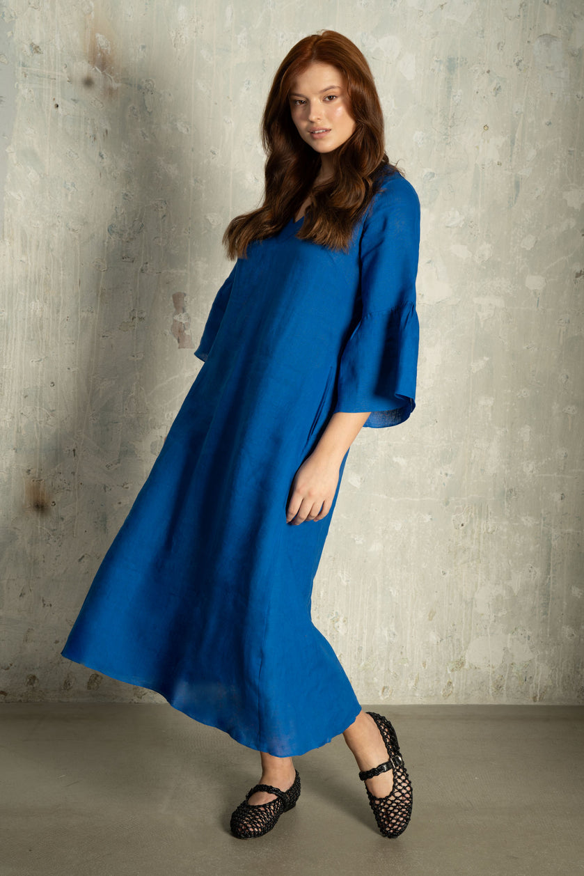 A woman modelling a flowing royal blue  linen midi dress with wide three-quarter sleeves and black lace-up flats, standing against a textured concrete wall.